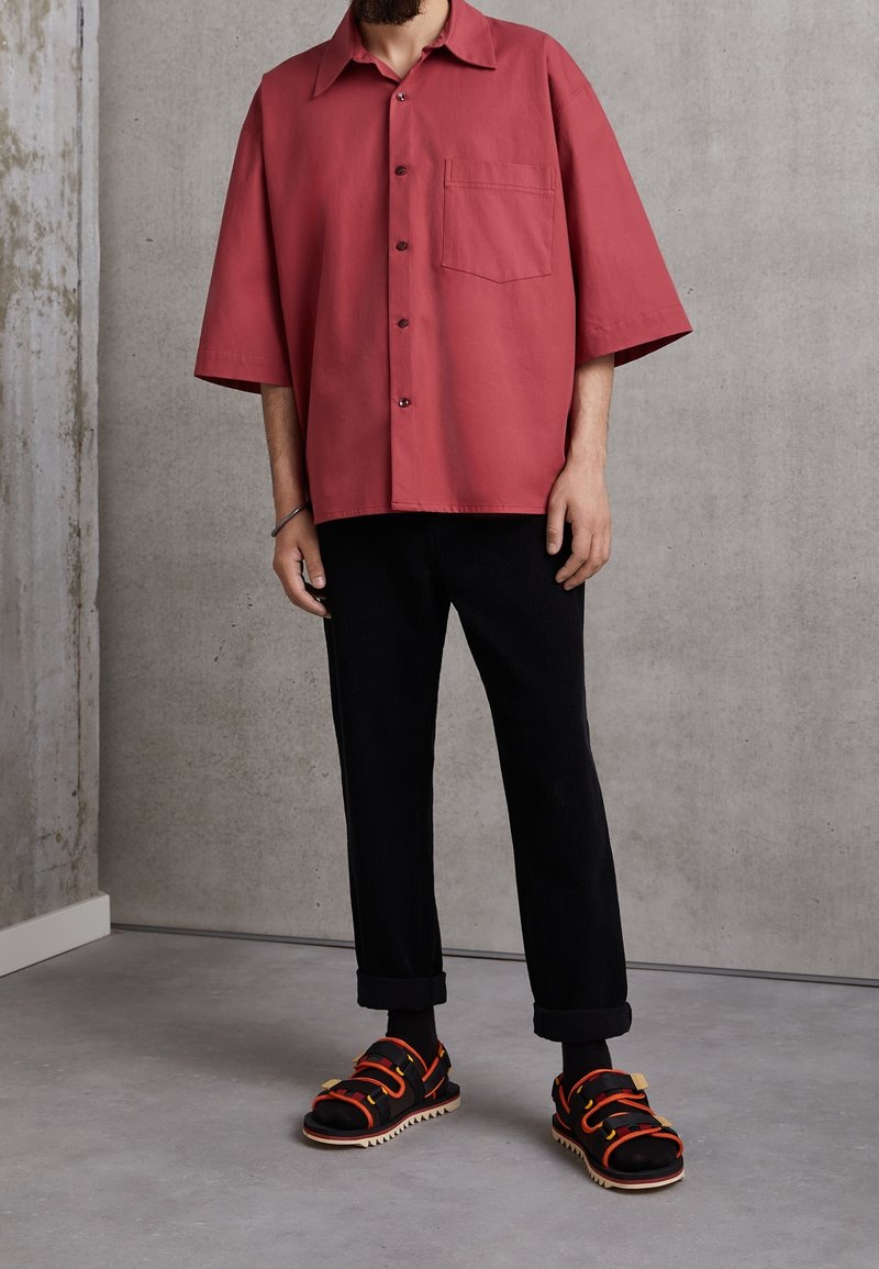 Košile s krátkým rukávem na knoflíky v barvě prachové růže, s jednou náprsní kapsou. Kombinováno s černými manšestrovými kalhotami a masivními černými sandály.