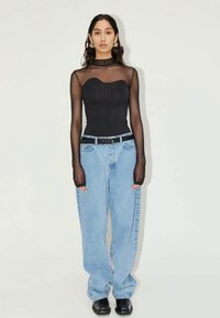 Woman wearing sheer black long-sleeve top with black bandeau, loose light blue jeans, black belt, and black shoes, standing against plain background.