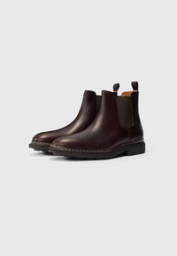 Bottines Chelsea en cuir marron foncé avec des panneaux élastiqués, des languettes de tirage et des semelles en caoutchouc cousues sur un fond clair.