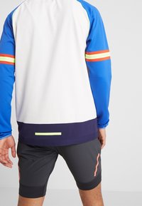 Lekka koszulka sportowa w kolorze białym, niebieskie rękawy z czerwonymi i żółtymi paskami. Do tego ciemne spodenki z delikatnym detalem w postaci brzoskwiniowej linii.