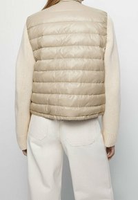Beżowa kamizelka puchowa z poziomym pikowaniem, wysokim kołnierzem i lekkim materiałem. Nosi się ją na kremowym swetrze, w połączeniu z jasnymi spodniami.