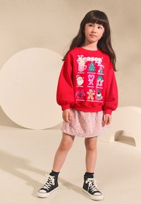 Felpa rossa con grafiche festive, abbinata a una gonna rosa scintillante. Scarpe da ginnastica nere alte completano il look.