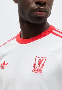 Artimas žmogaus, vilkinčio baltą Liverpulio futbolo klubo marškinėlį su raudonu apykaklės ir emblema kairėje krūtinės pusėje, vaizdas.