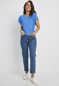 Niebieska koszulka z krótkim rękawem w białe kropki, zestawiona z jasnoniebieskimi dżinsami i białymi butami wsuwanymi, stojąca na jednolitym tle.