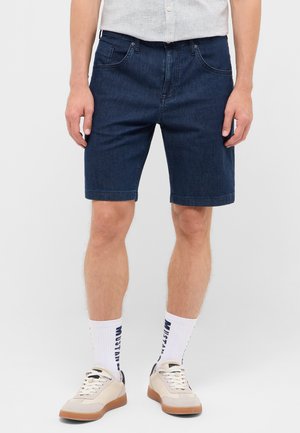 Mann trägt dunkelblaue Jeansshorts, weiße Socken mit Text, cremefarbene Sneaker und ein hellgraues Hemd.