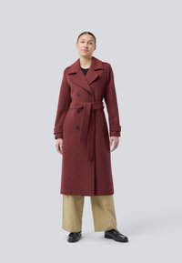 Kvinna iklädd en lång burgundy belagd kappa över beige vida byxor och svarta loafers, ståendes mot en enkel ljusgrå bakgrund.