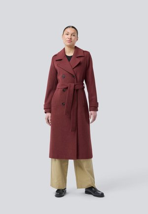 Mulher vestindo um longo casaco burgundy duplo com cinto, calças largas bejes e mocassins pretos, de pé contra um fundo cinza claro liso.