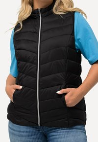 Svart pufferväst med dragkedja fram, horisontell quiltning och sidofickor, tillsammans med en ljusblå skjorta och jeans.