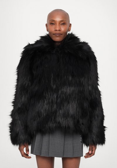 Veste en fausse fourrure noire avec un col haut, des manches longues et une finition texturée. Portée sur une jupe grise à rayures.