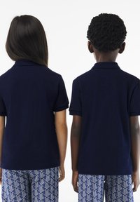 Dwoje dzieci w granatowych polo z krótkimi rękawami, pokazanych od tyłu. Dziewczynka ma długie włosy, a chłopiec ma krótkie, teksturowane włosy.