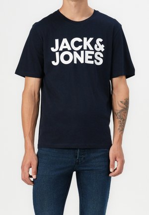 Mann trägt ein schwarzes kurzärmeliges T-Shirt mit weißem "JACK & JONES"-Schriftzug, dazu blaue Jeans und ein Tattoo auf seinem linken Unterarm.