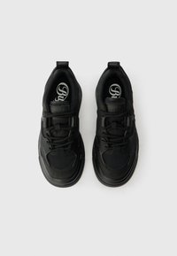 O pereche de pantofi sport negri cu șireturi, cu tălpi groase, logo pe tălpile interioare și benzi de prindere pe călcâie, expuși pe un fundal alb.