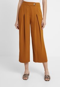 Kvinna som bär bruna högmidjade, vida cropped byxor med veck och knappdetalj, i kombination med sandaler med leopardmönster.