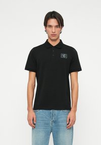 Černé polo tričko vyrobené z bavlny, se slim fit st�řihem, límečkem a nášivkou na hrudi. Kombinováno se světle modrými džíny.