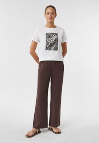 T-shirt blanc à manches courtes avec un imprimé graphique en noir et blanc, associé à un pantalon lâche marron et des sandales plates à lanières.