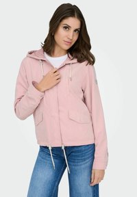 Femme portant une veste à capuche rose clair avec des cordons blancs, un jean bleu et une chemise blanche, posant avec une main près de la fermeture éclair de la veste.