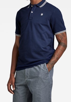 Homme portant un polo bleu marine avec liseré blanc et logo, pantalon gris clair rayé, tatouages visibles sur le bras droit, main droite dans la poche.