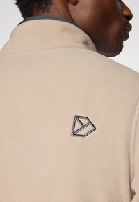 Giacca in fleece beige con colletto alto, caratterizzata da un logo ricamato blu scuro sul retro. Texture liscia e piena visibilità del logo.