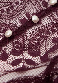 Burgundy lace fabric featuring intricate floral patterns, embellished with three shiny pearl buttons along the center.