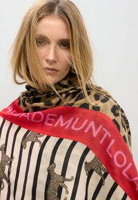 Bufanda de estampado de leopardo con un fondo beige, rayas negras y un borde rojo vibrante con texto. Fabricada en un material suave y ligero.