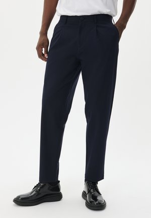 Pantaloni eleganti blu navy realizzati in tessuto morbido, con frontale plissettato, tasche inclinate e chiusura con bottone, abbinati a scarpe nere.