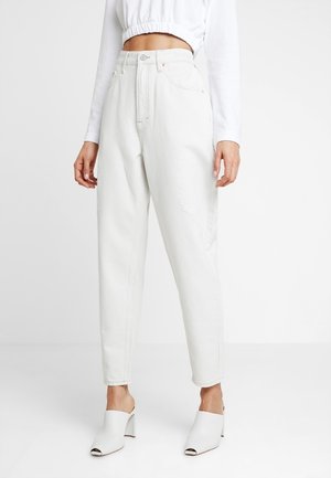 Femme portant un jean blanc taille haute, des mules blanches ouvertes à l'avant avec talons, et un top blanc court à manches longues, debout sur un fond blanc.
