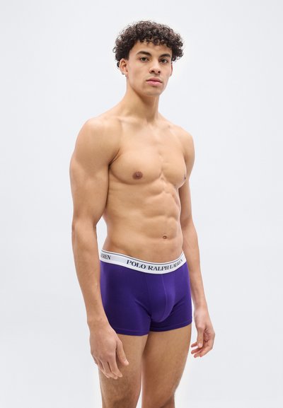 Νεαρός μυώδης άνδρας που στέκεται, φοράει μωβ boxer briefs Polo Ralph Lauren με λευκή ζώνη μέσης σε απλό λευκό φόντο.