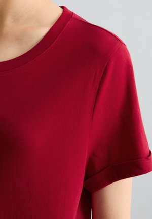 Close-up van een persoon die een effen rood T-shirt met korte mouwen en een ronde hals draagt tegen een neutrale achtergrond.