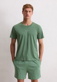Blond man wearing a green Marc O'Polo t-shirt and matching shorts, standing with hands in pockets against a plain white wall.