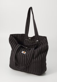 Bolso de lona negro con rayas verticales blancas, dos asas largas y cierre a presión. Presenta un pequeño parche de logo en la parte delantera.