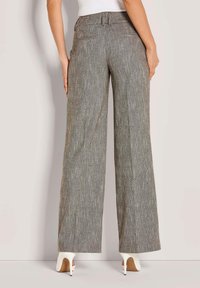 Vrouw in hoge taille, wijde grijze broek en witte hoge hakken, staand tegen een effen lichte achtergrond.