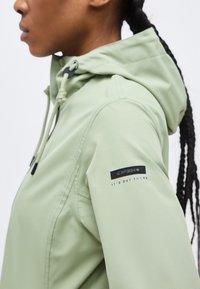 Γυναίκα με πλεξούδες που φοράει ανοιχτοπράσινο μπουφάν με κουκούλα, με το λογότυπο "ICEPEAK" και το κείμενο "IT'S OUT THERE" στο αριστερό μανίκι.