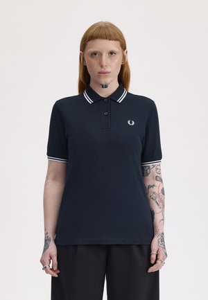 Jeune personne aux cheveux roux et tatouages, portant un polo bleu marine avec un col blanc rayé et un pantalon noir, debout devant un fond uni.