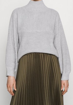 Personne portant un pull à col roulé en maille gris clair et une jupe plissée vert olive, debout devant un fond uni.