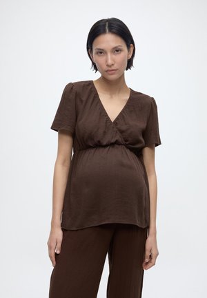 Ciążowa kobieta z krótkimi ciemnymi włosami, ubrana w brązową bluzkę z dekoltem w serek i pasujące spodnie, stojąca na jednolitym białym tle.