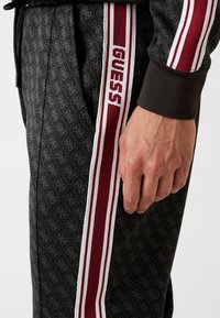 Melnas sportiskās bikses ar maroon un baltām vertikālām svītrām un "GUESS" logo sānos, papildinātas ar ribinātiem apkaklēm un jostu.