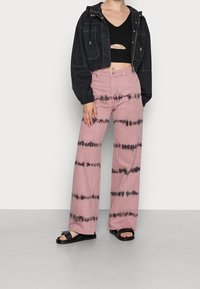 Avkortad svart jacka över en svart crop top, kombinerad med ljusrosa vida byxor med svart tie-dye mönster och svarta sandaler.