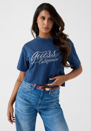 Donna con capelli lunghi e mossi che indossa una maglietta corta blu Guess California e jeans blu a vita alta con cintura rossa, posando con la mano sul fianco.