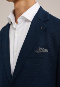 Blazer blu navy con texture e rever a punta, indossato sopra una camicia bianca, con un fazzoletto da taschino a motivo in toni di grigio.