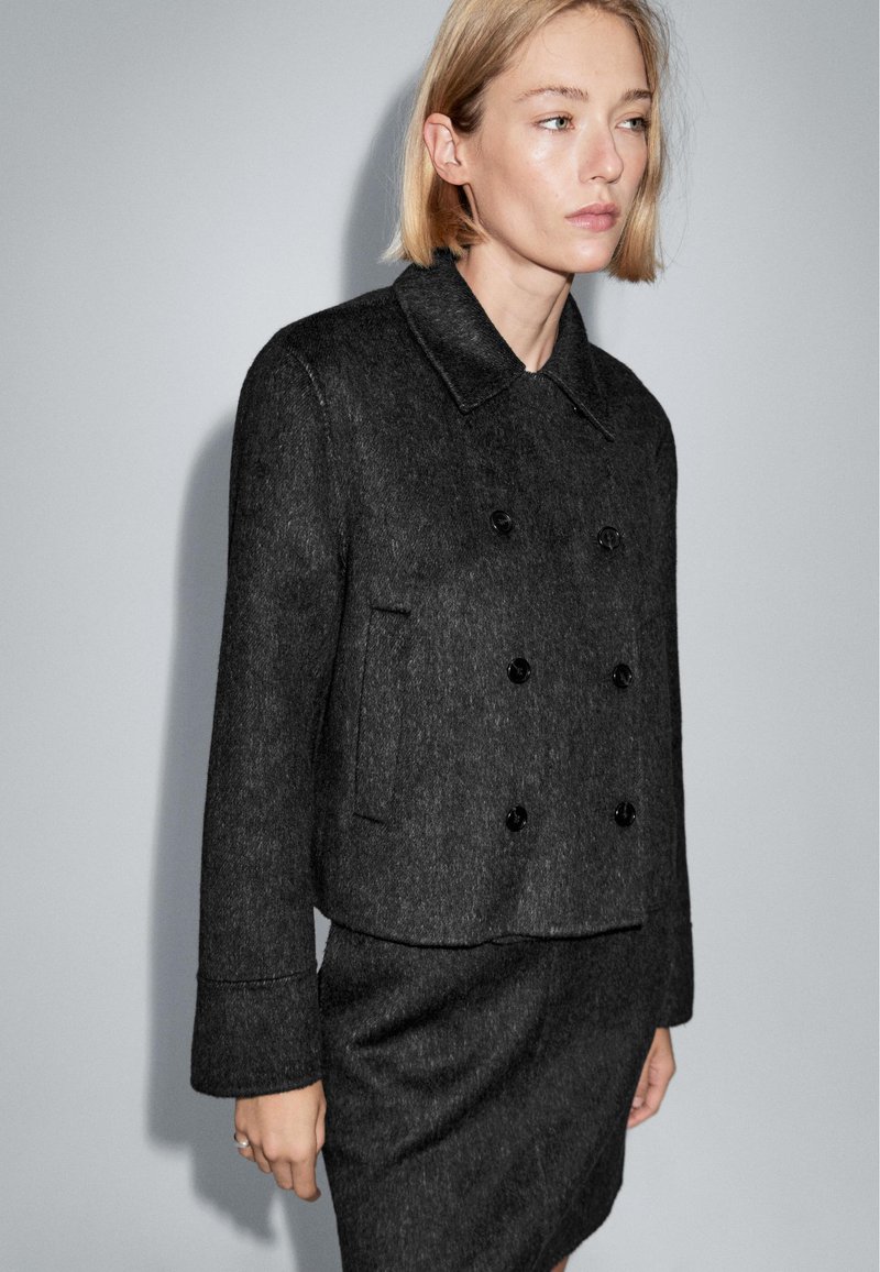 Kvinde med kort, blond hår iført en mørkegrå dobbeltradet uldjakke og matchende nederdel foran en ensfarvet grå baggrund.