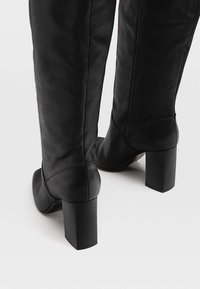 Bottes en cuir noires jusqu'au genou avec de gros talons carrés, présentées de dos sur un fond gris clair.