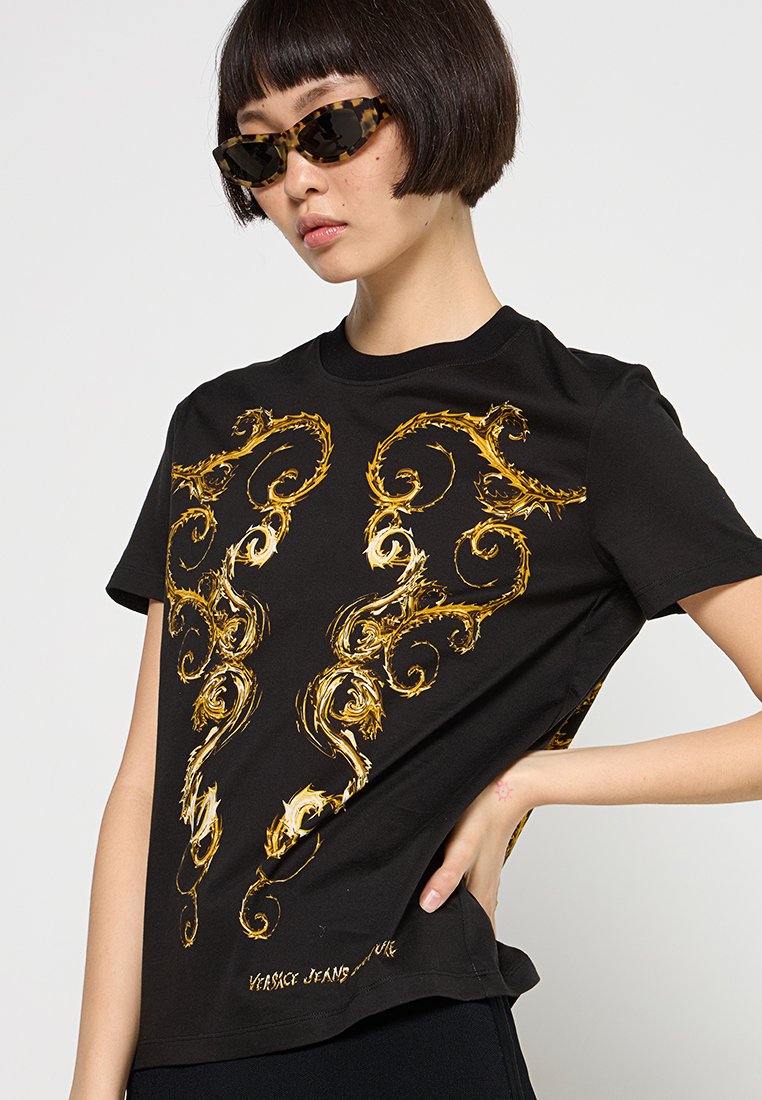 T-shirt noir avec des motifs baroques dorés en spirale. Manches courtes, col rond et coupe ample. Le texte de la marque au bas indique "Versace Jeans Couture."
