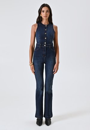 Salopette in denim scuro con collo alto, chiusura frontale con bottoni, vita aderente e gambe svasate. Cuciture a contrasto evidenti e colore blu classico.