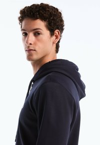 Sweat à capuche bleu marine en tissu texturé, avec une poche avant et une fermeture éclair. Comprend des cordons de serrage au niveau de la capuche.