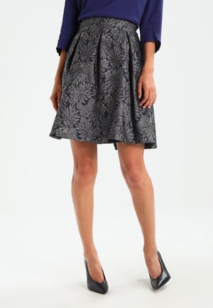 Vrouw met een knielange zwarte en zilveren rok met bloemmotief, een donkerblauw topje en zwarte puntige hoge hakken, staand op een witte achtergrond.