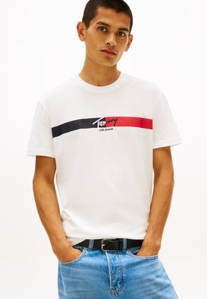 Νέος άνδρας που φοράει λευκό t-shirt Tommy Jeans με μαύρες και κόκκινες ρίγες και μπλε τζιν, στέκεται με τα χέρια στις τσέπες.