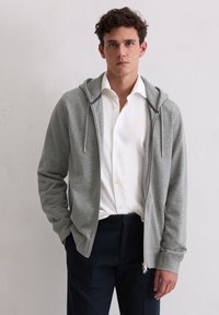 Felpa grigia con zip e cappuccio, caratterizzata da una superficie liscia, cordini regolabili e due tasche laterali. Indossata sopra una camicia bianca con bottoni.