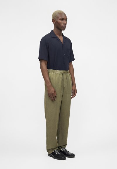 Άνδρας με κοντά ξανθά μαλλιά που φοράει μπλε σκούρο κοντομάνικο πουκάμισο, φαρδιά χακί παντελόνια και μαύρα δερμάτινα μοκασίνια, στέκεται πλαγίως.