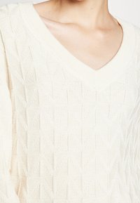 Pull à col en V en tricot de couleur crème, présentant un motif en losanges texturé et des manches larges et amples.