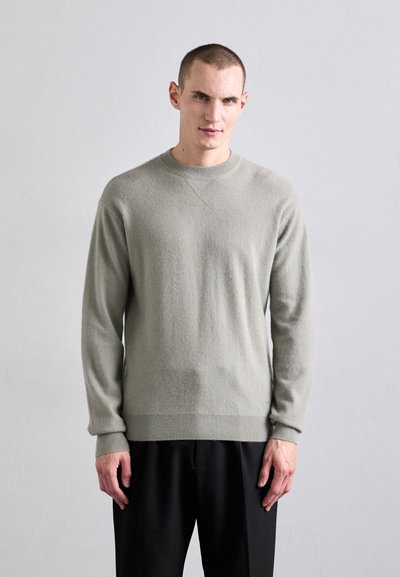 Pull en tricot gris clair avec un col rond, des manches longues et un ourlet côtelé, associé à un pantalon ample foncé.